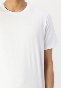 T-shirt en coton blanc avec un col rond, des manches courtes et un détail de logo discret sur la poitrine. Texture douce, coupe classique.