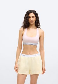 Γυναίκα με σγουρά μαλλιά που φοράει αθλητικό σουτιέν ανοιχτό ροζ και ανοιχτό κίτρινα σορτς με ζώνες μέσης με λογότυπο Calvin Klein, στέκεται μπροστά σε λευκό φόντο.