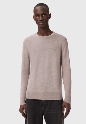 Mies, joka pukeutuneena vaalean beigeen pitkähihnaiseen neulepuseroon, jossa on pieni kirjailtu logo rinnassa, yhdistettynä tummiin farkkuihin.