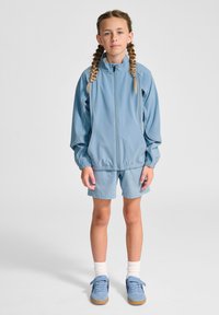 Ljusblått träningsset bestående av en jacka med dragkedja och elastiska muddar samt matchande shorts. Bärs med ljusa strumpor och blå sneakers.