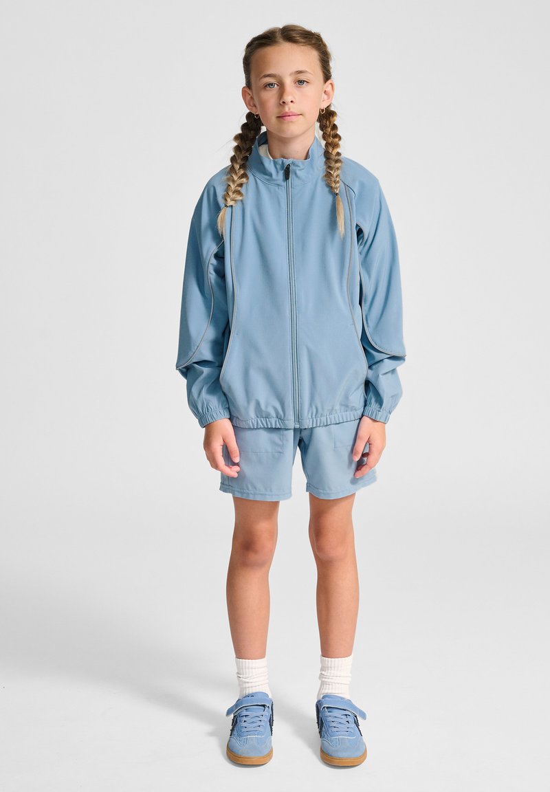 Ljusblått träningsset bestående av en jacka med dragkedja och elastiska muddar samt matchande shorts. Bärs med ljusa strumpor och blå sneakers.