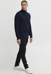 Pulover din tricot cu guler înalt de culoare bleumarin, cu un model texturat, asociat cu pantaloni negri slim-fit și sneakersi albi. Design minimalist fără logo-uri.
