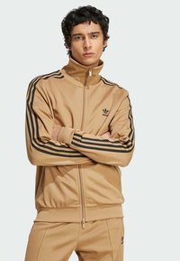 Tamni zip-up sportski jakna boje kaki s crnim trostrukim wergetama na rukavima i logom na prsima. Glatka tekstura, uski kroj, visoki ovratnik.