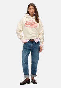 Crème hoodie met rood SuperDry-logo, gecombineerd met een lichtroze shirt, blauwe jeans met opgerolde mouwen en donkere loafer schoenen, die een casual stijl benadrukken.