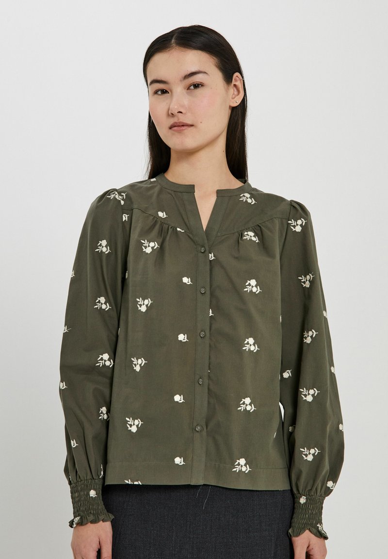 Olivgrön blus med vit blommönstring, knapplist fram, veckad detalj och smockade ärmslut. Mjuk bomullstyg.
