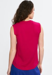 Fuksjowa bluzka bez rękawów z okrągłym dekoltem i zaokrąglonym dołem, wykonana z gładkiego materiału. Widok z tyłu ukazuje cięcie na ramionach i strukturę tkaniny.