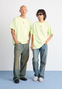 Světle zelené oversized trička s logem, spárované s volnými modrými džíny. Modeli nosí sluneční brýle a pohodlnou obuv. Hladká textilie.