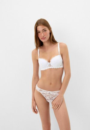 Young woman with long brown hair wearing white lace bra and matching lace underwear standing against plain white background.