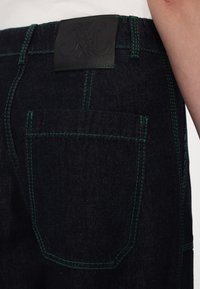 Donkerblauwe denim jeans met groene stiksel, een zwart leren label op de achterkant van de tailleband, en een achterzak met gedetailleerd stiksel.