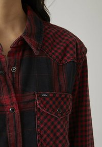 Chemise en flanelle à carreaux rouges et noirs avec un col à boutons, une fermeture à pression et une poche poitrine arborant une petite étiquette logo.