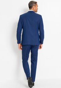 Completo blu navy con design strutturato, taglio dritto e texture liscia. Presenta pantaloni su misura e scarpe scure. Vista posteriore.