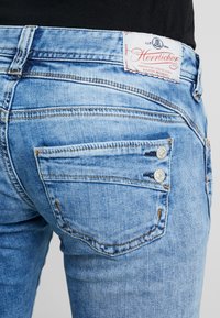 Ljusblå denimjeans med en detalj av tvåknappar på bakfickan och sydda accenter. Midjebandet har en logotypetikett.