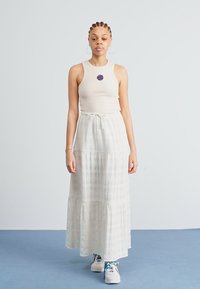 Top scurt crem asortat cu o fustă maxi albă, cu volane, având un model texturat. Modelul stă pe un fundal albastru, purtând teniși.