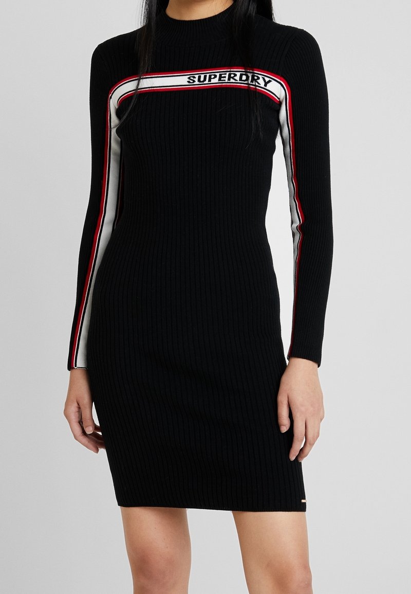 Robe noire à manches longues côtelées avec des rayures blanches et rouges traversant la poitrine et les manches, avec le texte "SUPERDRY" sur la bande de poitrine.