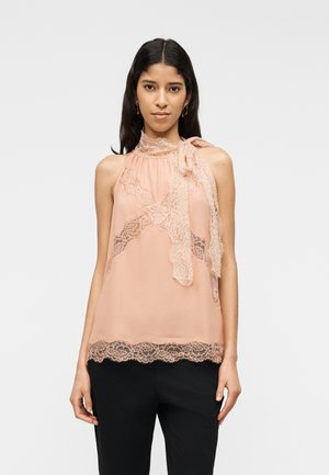 Vrouw draagt een mouwloze perzikkleurige kanten blouse met strik bij de hals en zwarte broek, staand tegen een effen lichte achtergrond.