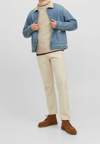 Jeansjacke mit hellblauem Waschung und hellbraunem Kragen, beiger Strickpullover, cremefarbene Hose und braune Wildlederschuhe. Das Model posiert mit den Händen in der Jacke.