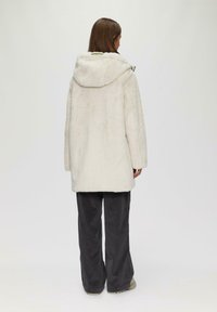 Manteau en fausse fourrure blanche avec capuche, présentant des manches longues, une coupe droite et une texture douce. Porté sur un pantalon en velours côtelé gris et des chaussures beiges.