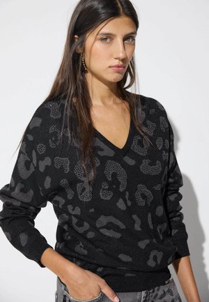 Mujer con el pelo largo y oscuro, vistiendo un suéter negro con cuello en V y un sutil estampado de leopardo, y vaqueros gris claro, de pie con una mano en el bolsillo.