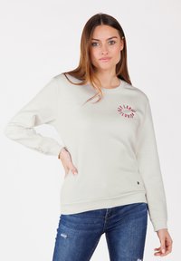 Felpa beige chiaro con maniche lunghe, scollo rotondo, stampa frontale rossa, polsini e orlo a coste; abbinata a jeans blu.