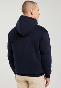 Vyras, stovintis nugara, dėvintis tamsiai mėlynį gobtuvinį džemperį ir smėlio spalvos kelnes, priešais vientisą šviesų foną.