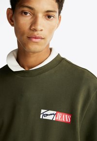 Sudadera de color verde oliva con un collar blanco, que presenta un parche rectangular de logo en rojo, blanco y azul, mostrando "Tommy JEANS."