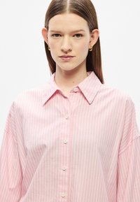 Camisa de botões às riscas rosa-claro, com gola clássica e corte folgado. O tecido parece macio com um brilho subtil; apresenta botões brancos.