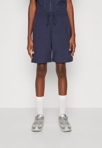 Marineblauwe shorts met een elastische tailleband, een trekkoord, zijzakken en lichte stikdetails, gecombineerd met witte sokken en zilveren sneakers.