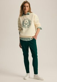 Kremowa bluza z graficznym motywem tenisa w kolorze zielonym, zestawiona z zieloną koszulą w paski pod spodem oraz ciemnozielonymi spodniami typu slim fit. Białe sneakersy.