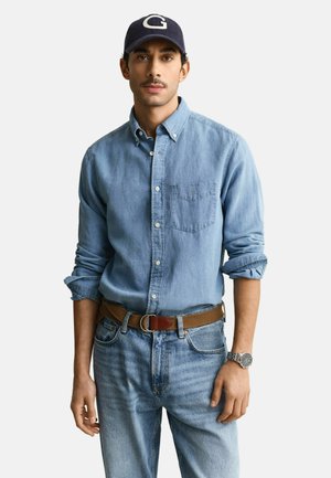 Homme portant une chemise en jean bleue aux manches retroussées, un jean bleu clair, une ceinture marron, une montre-bracelet argentée et une casquette bleu marine avec un logo "G" blanc.