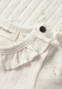 Vêtement pour bébé blanc doux avec petits motifs floraux, col à volants et bouton-pression noir près de l'encolure ; étiquette indiquant la taille 1-2 mois.