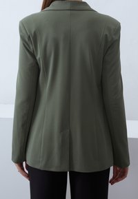 Olivově zelený ušitý blazer s dlouhými rukávy, klopovým límcem a rozparkem na zadní straně. Vyroben z hladké látky, s elegantním, strukturovaným siluetou.