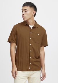 Hombre vistiendo una camisa de botones de manga corta marrón con botones blancos y pantalones beige, mirando hacia su izquierda contra un fondo blanco.