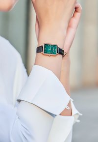 Grön fyrkantig klocka med guldaccent och svart läderarmband. Hand som vilar på en vit skjorta med uppvikt ärm. Enkla arabiska siffror.