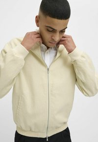 Veste zippée beige clair en tissu texturé, avec une taille élastique et de discrètes poches latérales, portée avec une chemise blanche à col en dessous.