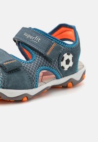 Blå og grå sandaler laget av stoff og gummi, med borrelåser, oransje detaljer og en fotballdetalj på siden.