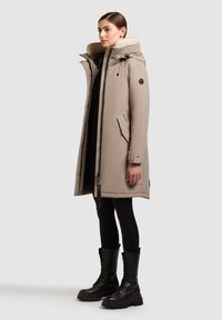 Parka imbottita beige con cappuccio foderato in pelliccia, dotata di zip, tasche laterali e un patch con logo. Stivali da combattimento neri con dettaglio delle stringhe.