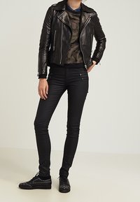 Černá kožená motorkářská bunda se stříbrnými zipy, pod ní maskáčová košile, přiléhavé černé džíny a hroty zdobené černé boty.