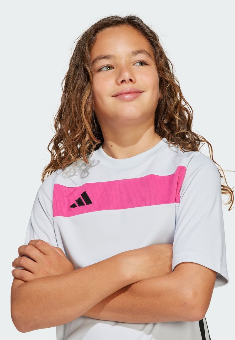 Majica za mlade sportiste u bijeloj boji s ružičastom horizontalnom prugom. Sadrži crni logotip i rebrasti uzorak na prugi. Kratki rukavi, opušteni kroj.