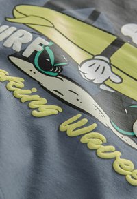 Tessuto grigio con un design grafico di un personaggio dei cartoni animati che fa surf, con accenti verdi, testo bianco in grassetto e elementi testurizzati gialli.
