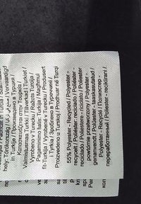 Vit tygetik med svart text som specificerar material: 55% återvunnen polyester. Fler språk vård- och produktionsinformation i olika typsnitt.