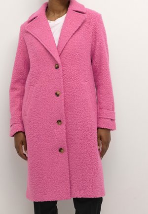 Vrouw draagt een knielange, gestructureerde roze jas met schildpaddenknopen over een witte top en zwarte broek.