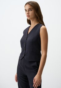 Gilet a righe blu navy con scollo a V profondo, chiusura a cinque bottoni e due tasche frontali; vestibilità su misura con una texture liscia.