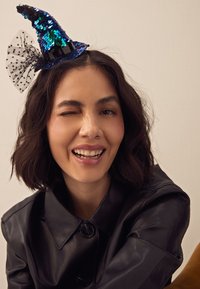 Sprankelende blauwe paillettenheksenhoed met een zwarte band en tule strik, gedragen door een persoon met donker haar op schouderlengte, in een zwarte leren jas.