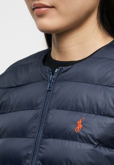 Κοντινό πλάνο ενός ατόμου που φοράει μπλε σκούρο μπουφάν με ραφές, με κόκκινο κεντημένο λογότυπο παίκτη πόλο στο στήθος και φερμουάρ μπροστά.