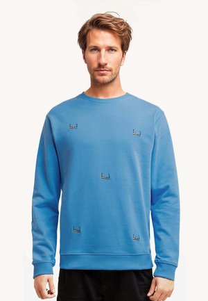 Młody mężczyzna o jasnobrązowych włosach, ubrany w niebieską bluzę z małymi wyszywanymi wzorami pływaków na przodzie oraz czarne spodnie.