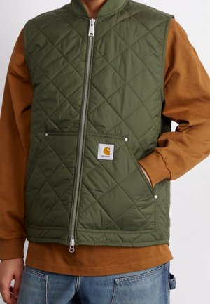 Personne portant une veste matelassée verte zippée sur une chemise marron à manches longues, une main dans la poche de la veste et un jean bleu.