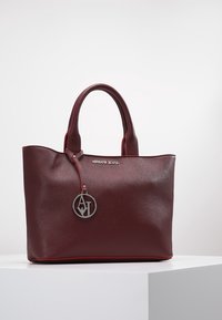 Vínově červená kabelka z texturované umělé kůže s kontrastním červeným prošitím, dvěma uchy a kulatým kovovým přívěskem s logem připevněným ke pásku.