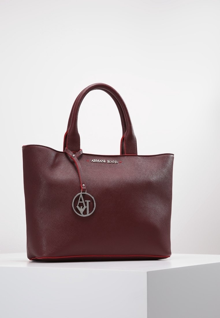 Tummanpunainen käsilaukku, jossa on teksturoitua keinonahkaa, kontrastivärisellä punaisella ompeleella, kaksi kahvaa ja pyöreä metallinen logovetokoru kiinnitettynä hihnaan.