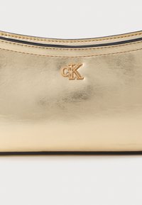 Χρυσό δερμάτινο πουγκί με λεία, ανακλαστική υφή. Διαθέτει μαύρο φερμουάρ και χρυσό λογότυπο "CK" ανάγλυφο μπροστά. Συμπαγής σχεδίαση.