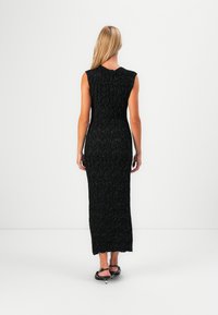 Vrouw met lang blond haar in een mouwloos, gestructureerd zwart maxi-jurk en zwarte strappy kitten heels, staande tegen een witte achtergrond.